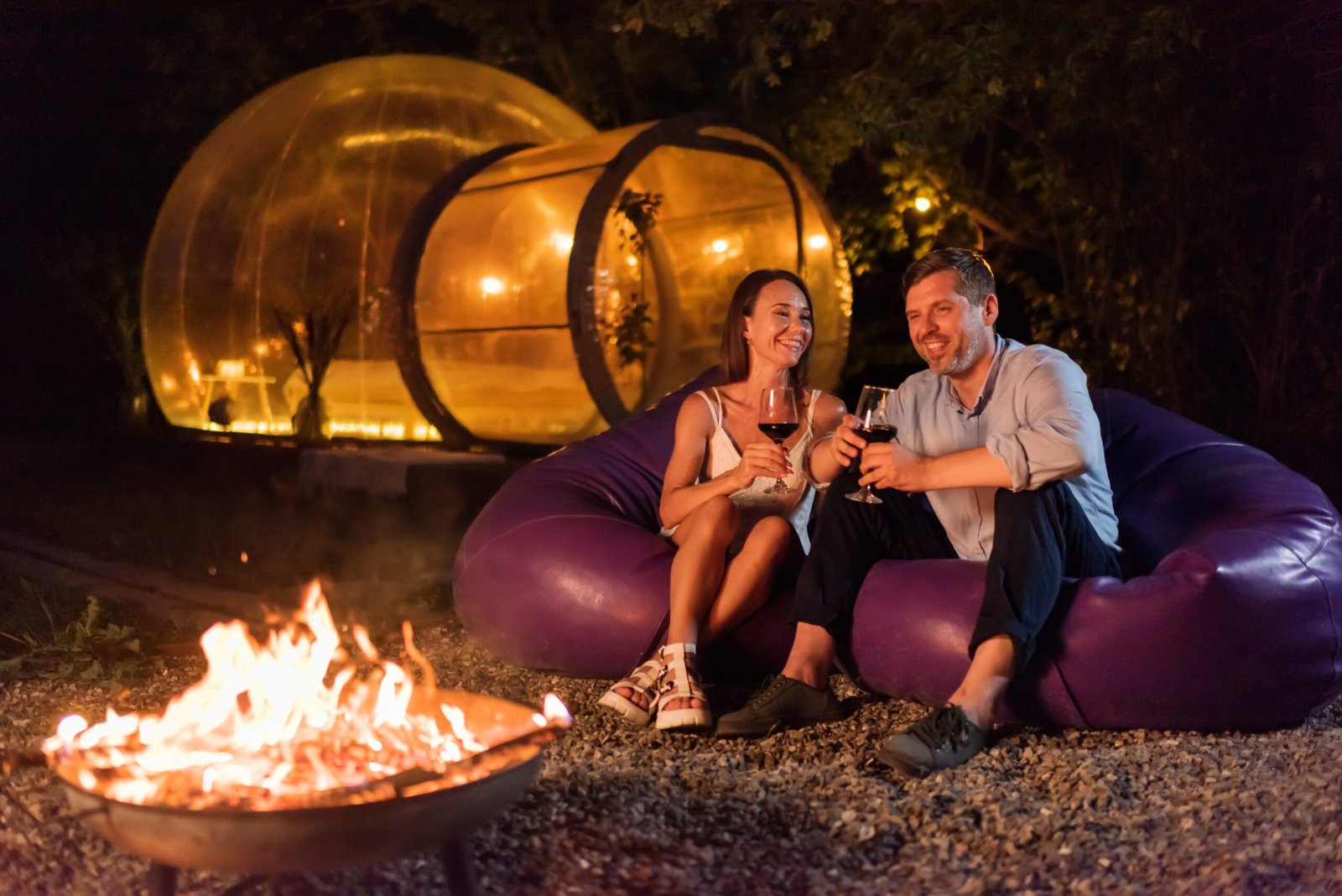 Pareja con una copa de vino junto a una chimenea dentro de una cúpula transparente, de noche