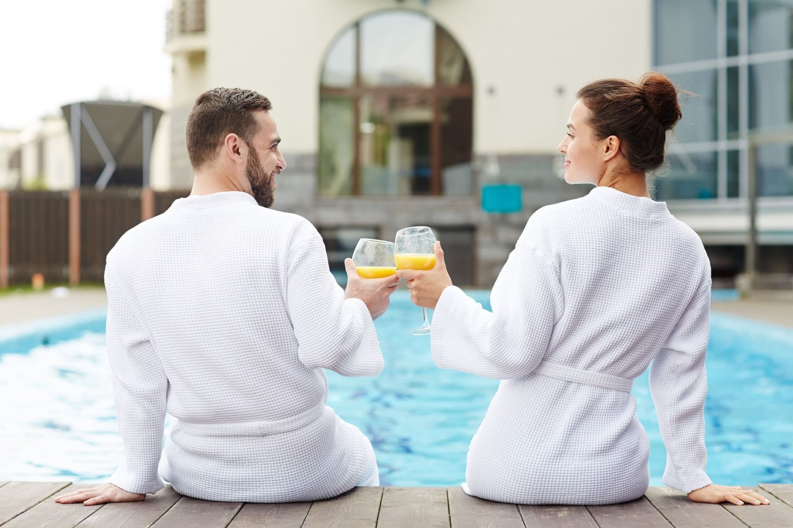 Pareja en albornoz brindando con zumo de naranja sentada en el borde de la piscina del hotel